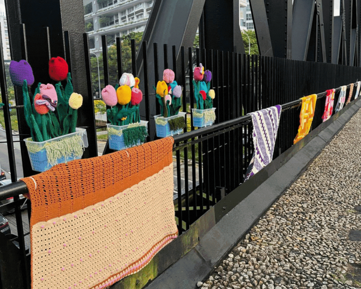 Crochet tulips in plastic pots and colourful knitted blankets displayed along a black metal bridge railing, with an urban streetscape behind.